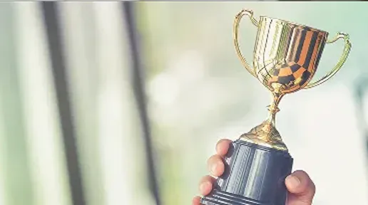 Hand holding a gold trophy cup with two handles against a blurred light background.