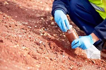 Gloved hands collecting a red soil sample into a jar