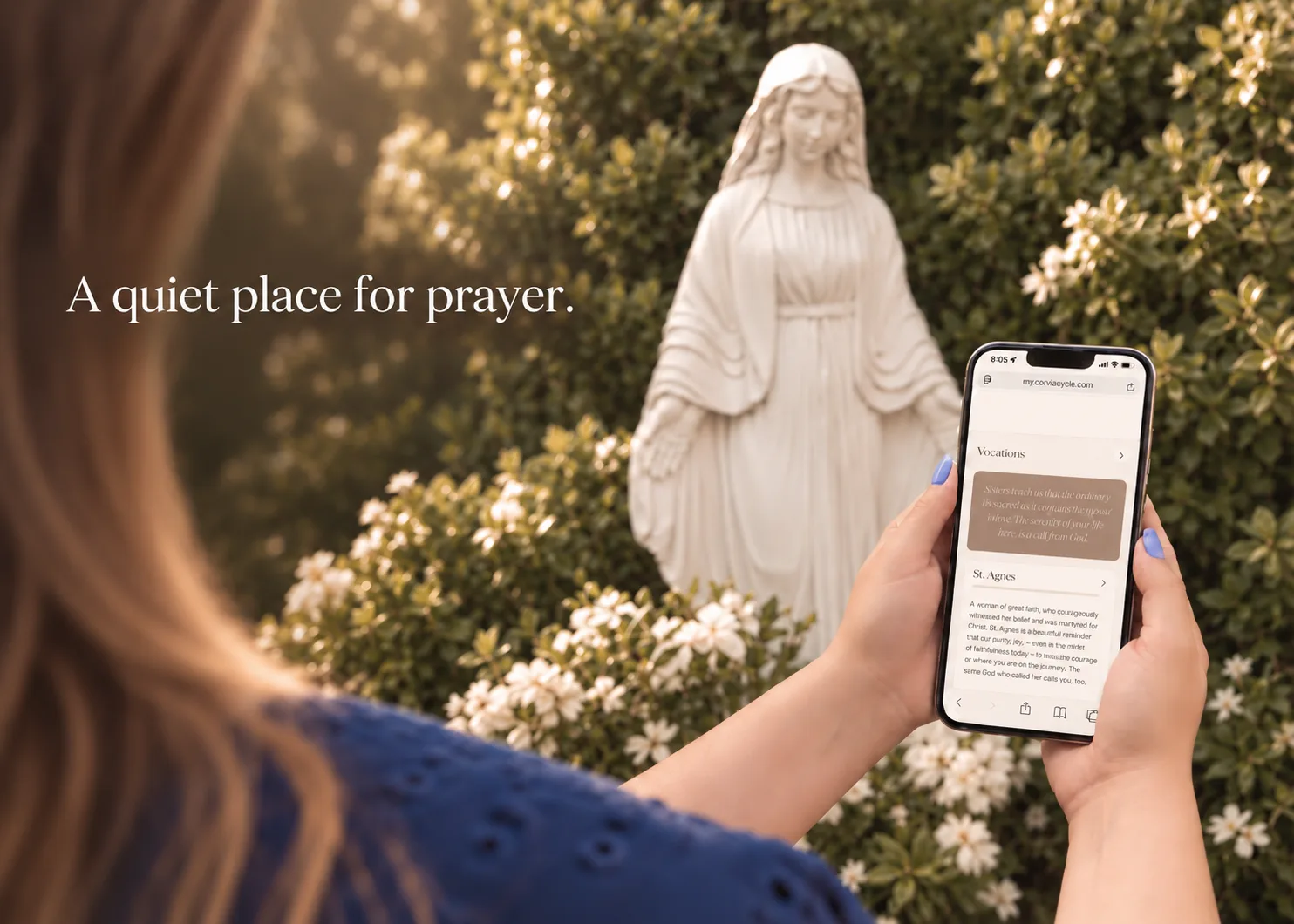 A woman holding her phone showing the Corvia prayer library, in front of a white Marian statue among flowering shrubs