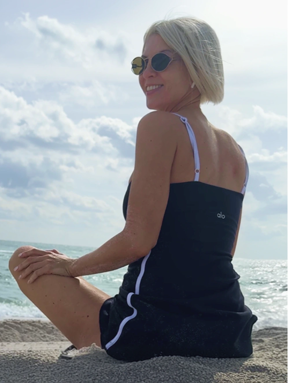 Woman with short blonde hair sitting on sandy beach facing ocean under cloudy sky, wearing sunglasses and black outfit.