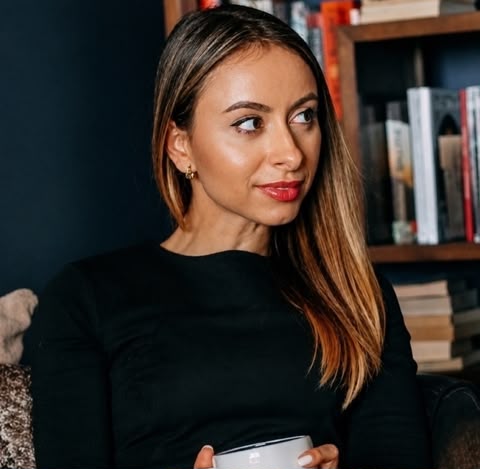 Woman with long hair in a black top holding a white cup, sitting near a bookcase with books.
