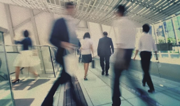 Blurred image of business people walking in a modern, bright office corridor with large windows and overhead architectural details.