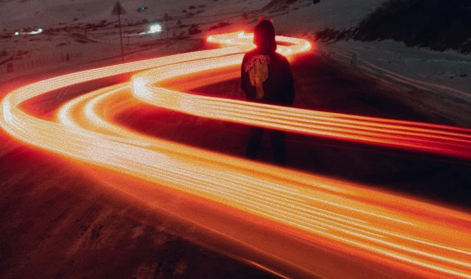 Person standing on a dark road at night with bright orange light trails swirling around them, creating a glowing path effect.