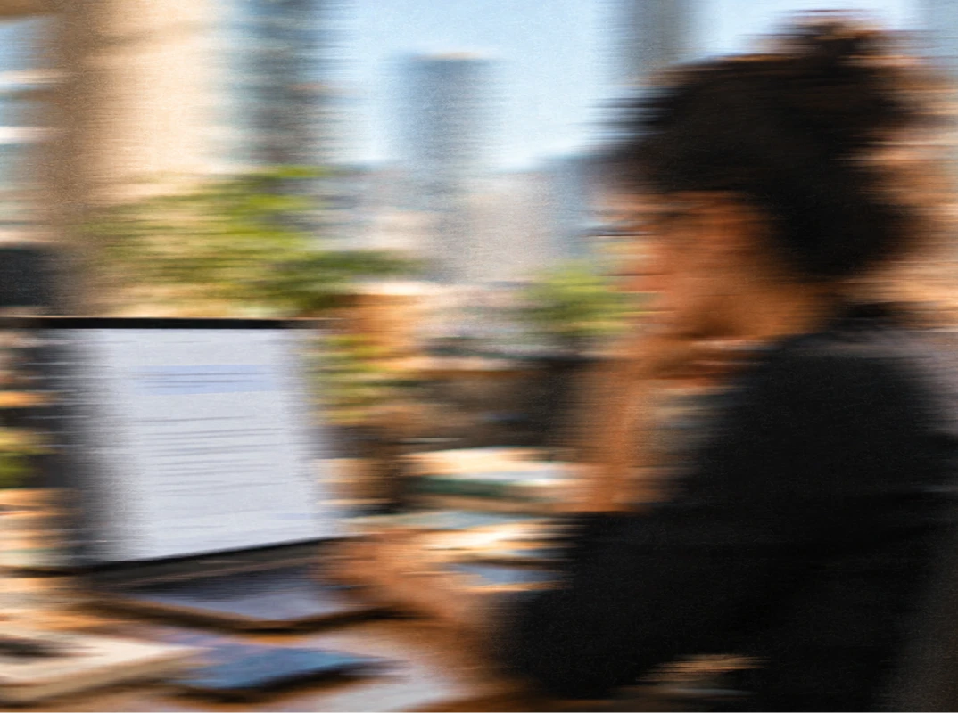 Blurred photo of a person working at a desk with a laptop displaying a document or webpage in an office setting.