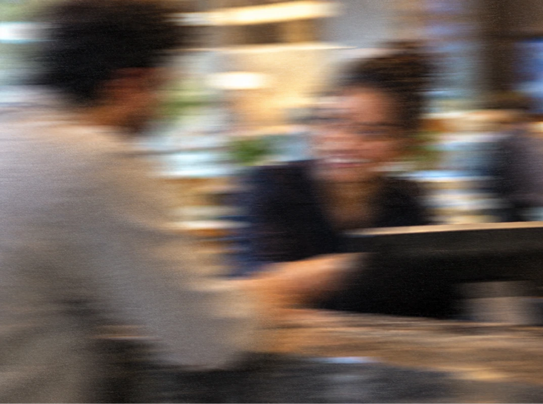 A blurred image showing two people in conversation across a table, with one person smiling and the background suggesting an indoor setting with warm lighting.