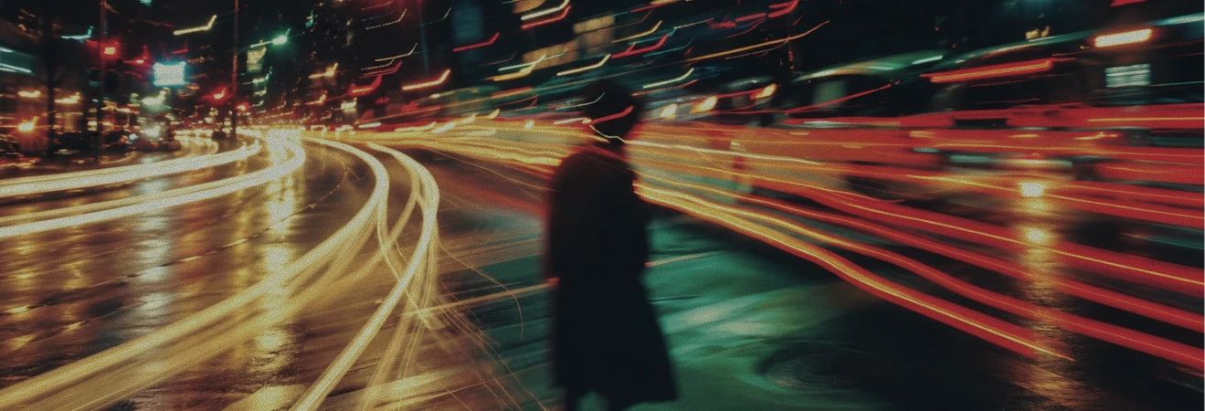 Blurred silhouette of a person walking on a wet city street at night with long exposure light trails from passing vehicles in white and red creating streaks across the scene.