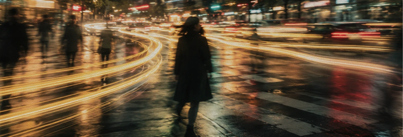 A blurred nighttime city street scene with light trails from moving vehicles and faint silhouettes of people walking on wet pavement.