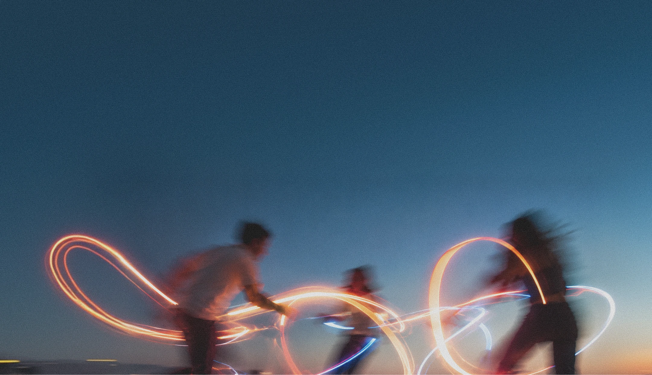 Three people in motion at dusk with colored light trails creating loops around them against a clear blue sky.