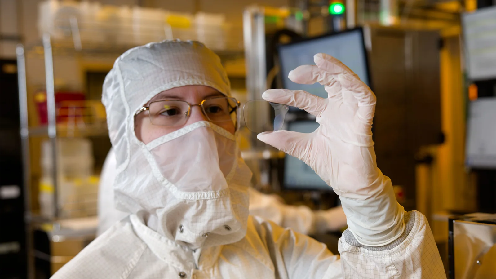 A Magic Leap employee in a full cleanroom coverall holding a small transparent waveguide lens up to the light inside a fabrication cleanroom