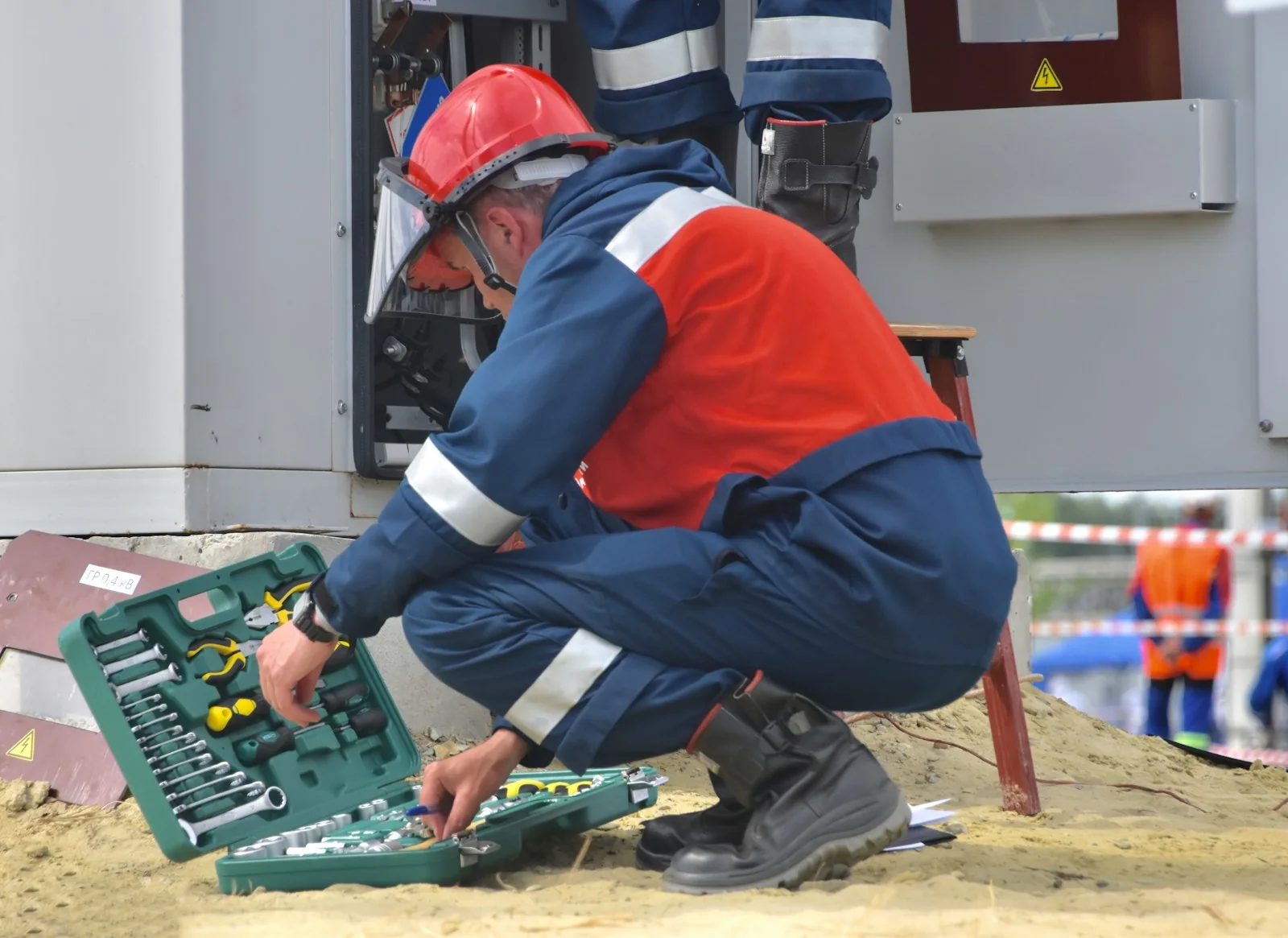 A technician in a red safety helmet and high-visibility overalls crouching at an open equipment cabinet on a work site, with an open socket set on the ground beside him