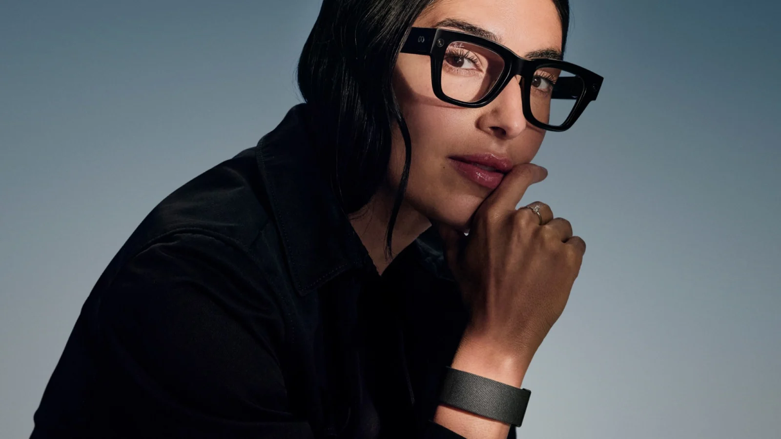 Person wearing black Meta Ray-Ban Display glasses with clear lenses and a small Meta logo on the temple arm, resting their chin on a hand that wears the fabric Meta Neural Band on the wrist