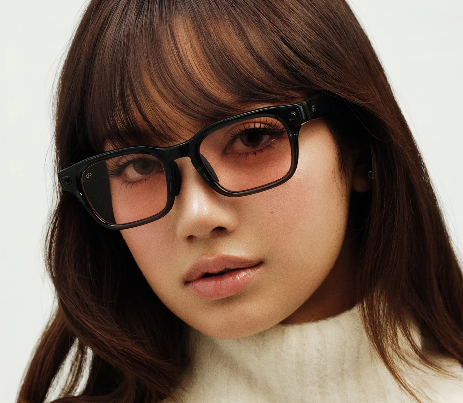Person wearing a dark rectangular Meta Glasses frame with blush tinted lenses, showing the camera at the right hinge and a small logo at the left hinge