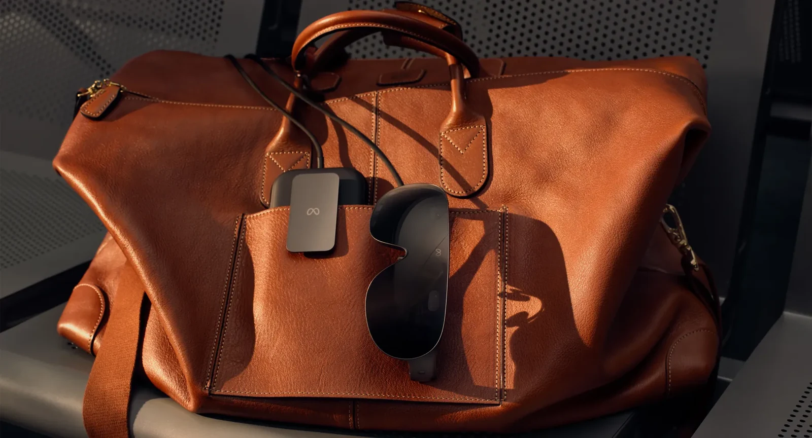 A tan leather holdall on a bench, with the Meta VR Glasses and their compute puck tucked into the outside pocket and the tether running between them