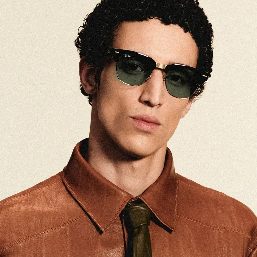 A person wearing Ray-Ban Meta Audio Clubmaster glasses in black and gold with green lenses, photographed against a pale background