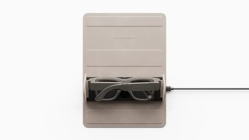 Meta Ray-Ban Display glasses folded inside a sand-colored foldable charging case with a charging cable running from it