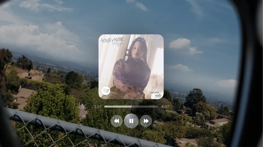 Meta's rendering of the view through Meta Ray-Ban Display, a music player card with album art and transport controls floating over a hillside view, framed by the edge of the lens