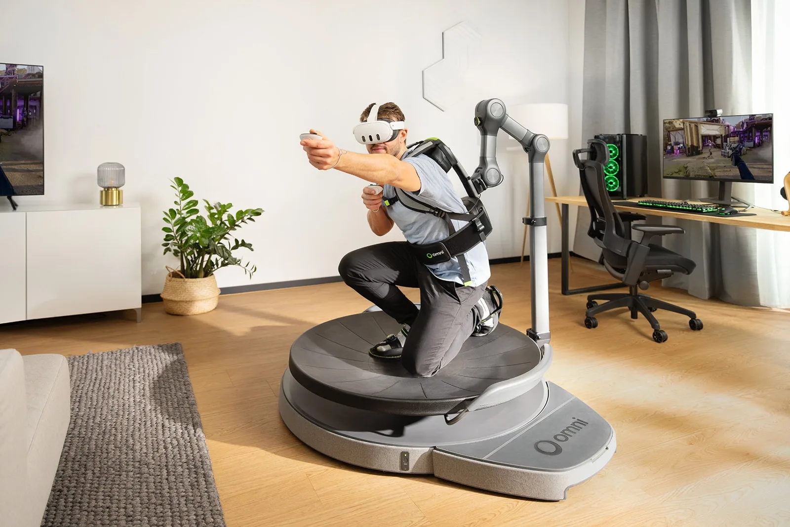 A player wearing a Meta Quest headset kneels on the circular base of the Virtuix Omni One treadmill, attached by the vest to the jointed support arm, aiming with Quest controllers in a living room