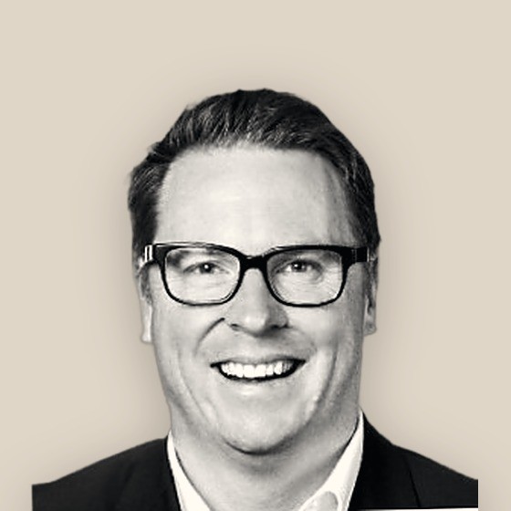 Anthony Spargo, smiling in a dark jacket, black-and-white on a warm sand background