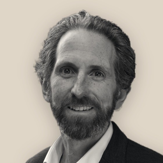 Tim Mahony, smiling in an open-collared shirt, black-and-white on a warm sand background