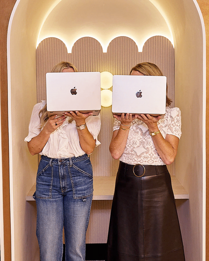 Sarah Hodgkins and Zoe White hold laptops over their faces, then lower them to reveal themselves smiling