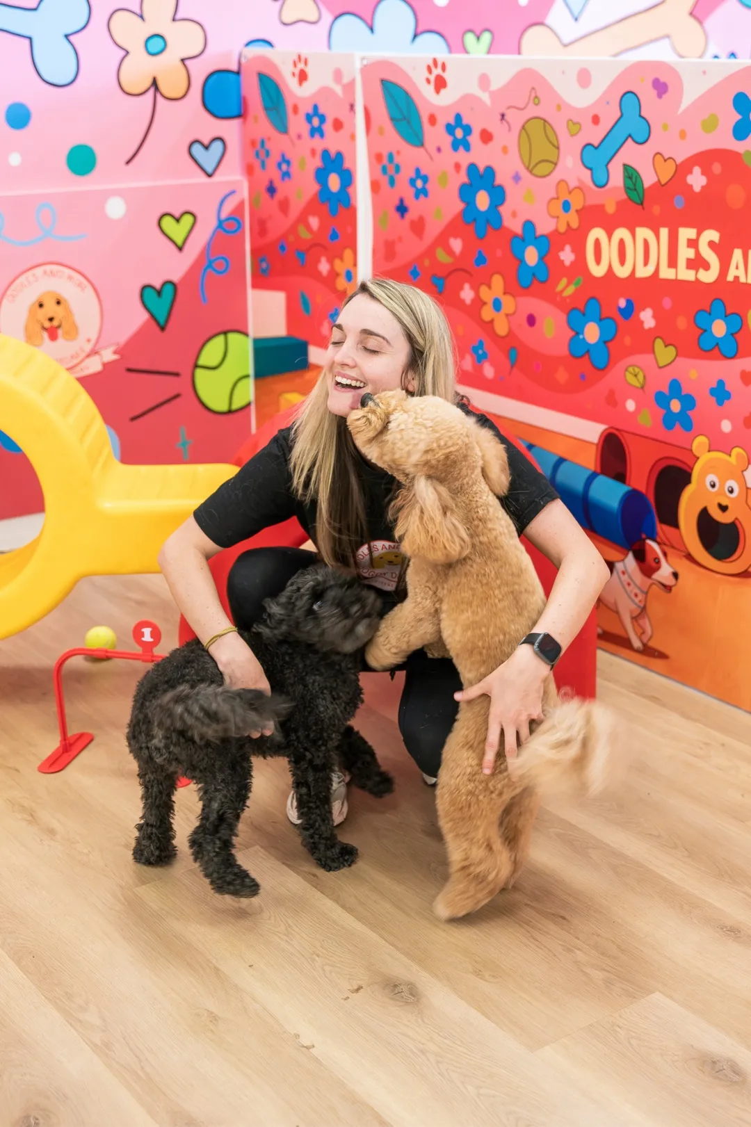 Woman smiling and crouching while two small dogs greet her in a colorful indoor play area.