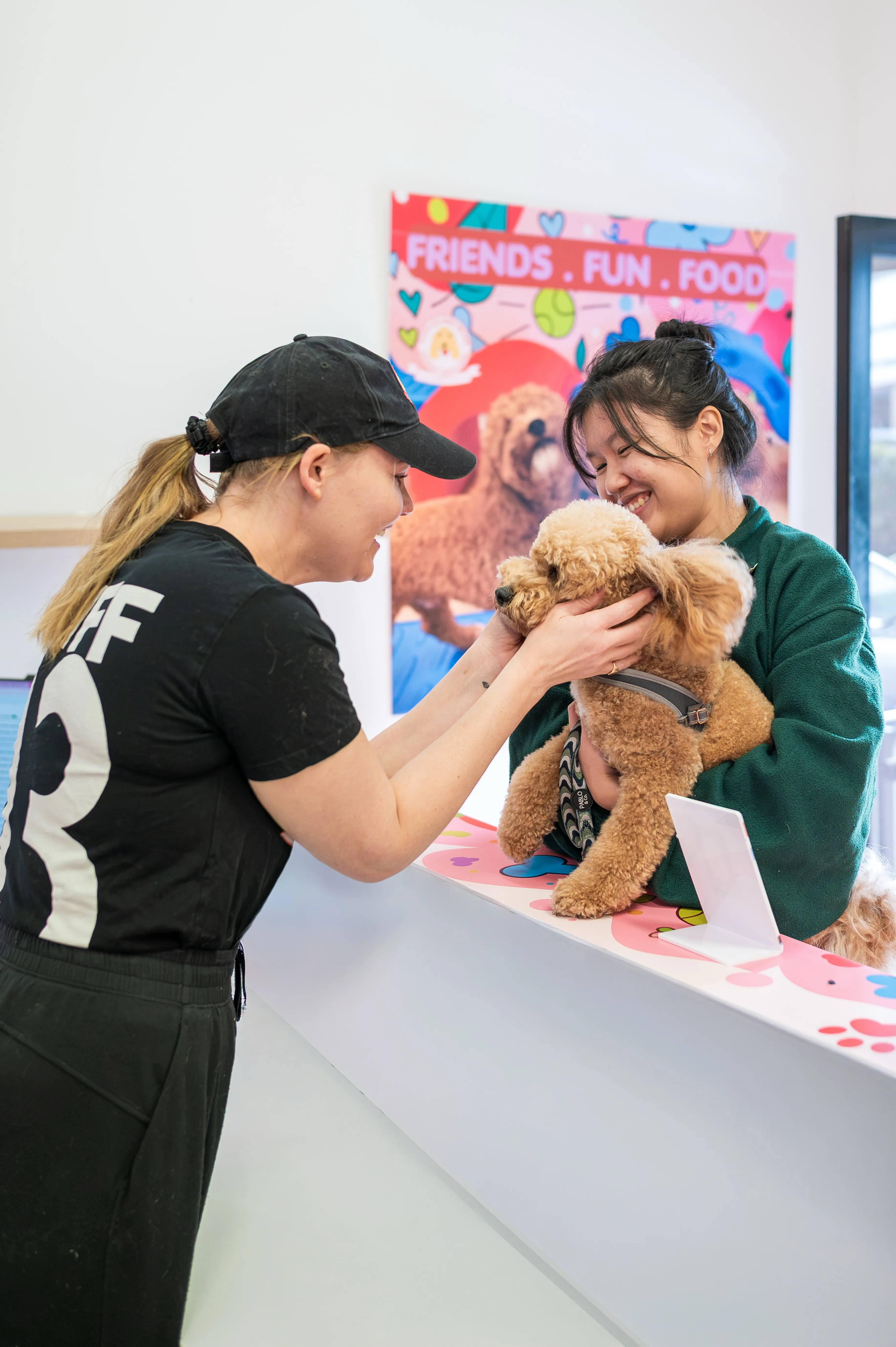 Two women smiling at a fluffy brown dog on a counter under a colorful Friends Fun Food sign.