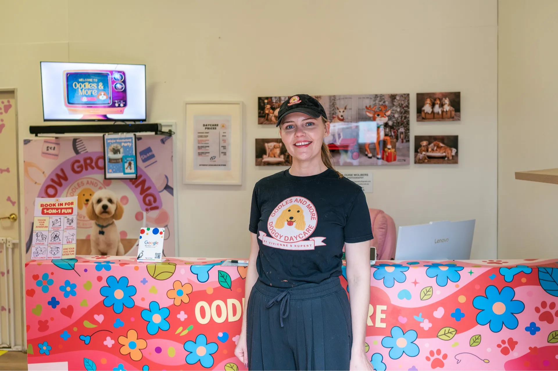 Smiling woman in black Oodles and More Doggy Daycare shirt stands behind colorful reception counter.