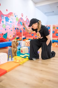 A woman in a black cap and outfit kneels on a wooden floor, raising her hand to train a small fluffy dog sitting attentively on colorful play mats in a bright room with playful mural walls.
