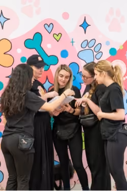 Five women in black outfits standing together indoors in front of a colorful wall with large paw prints, hearts, and bones; they are looking at a clipboard one of them is holding.