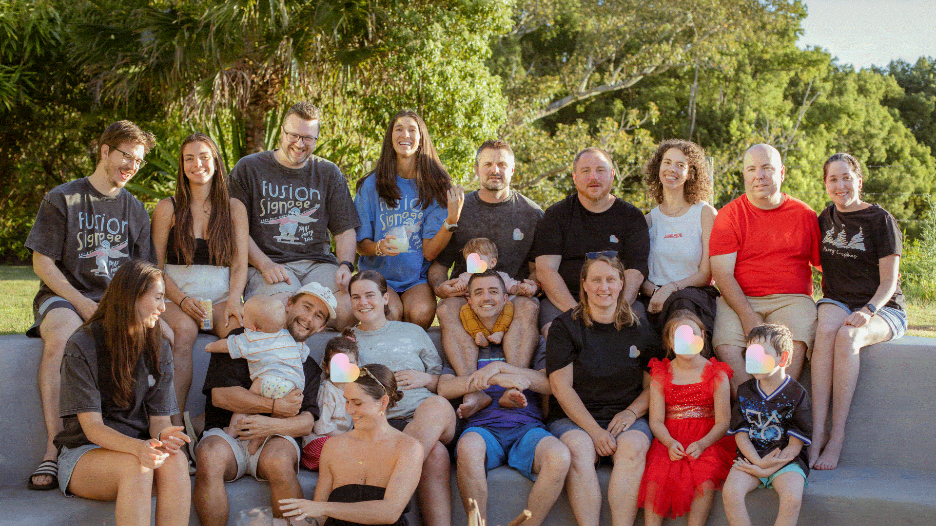 Group of adults and children sitting outdoors on steps, smiling and enjoying a sunny day in a green area.