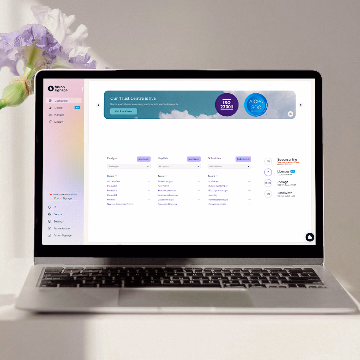 Laptop on a white surface showing a Fusion Signage dashboard with sections for Designs, Playlists, and Schedules, plus status indicators for screens online, licenses, storage, and bandwidth.