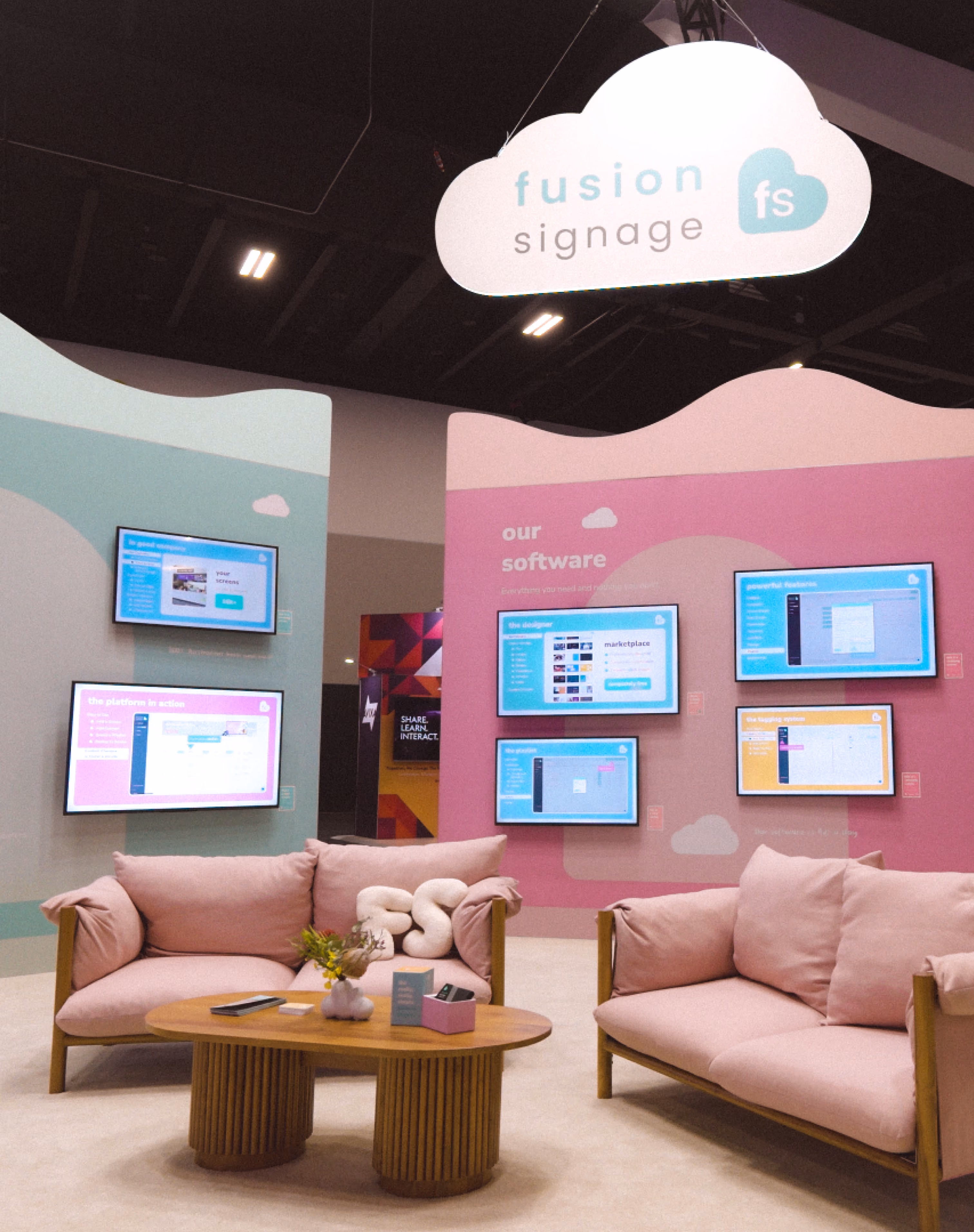 Exhibition booth with pastel pink and teal walls displaying Fusion Signage software on multiple screens, a white cloud-shaped ceiling sign with the logo, and two pink cushioned sofas around a wooden coffee table with a plant and promotional materials.