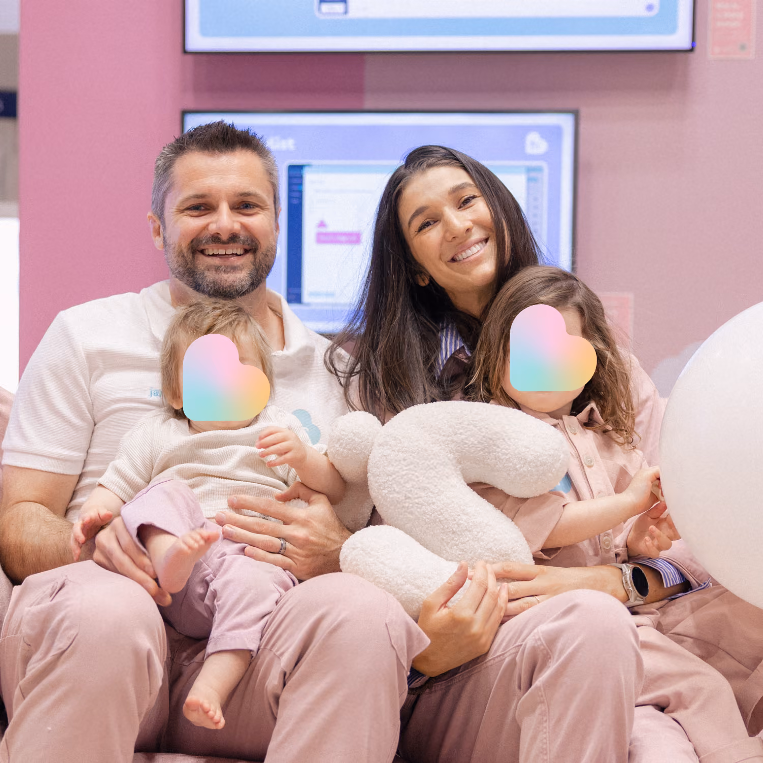 Smiling man and woman sitting closely together holding two small children with pastel heart stickers obscuring their faces, the woman holding a white plush letter S, against a pink and blue digital display background.