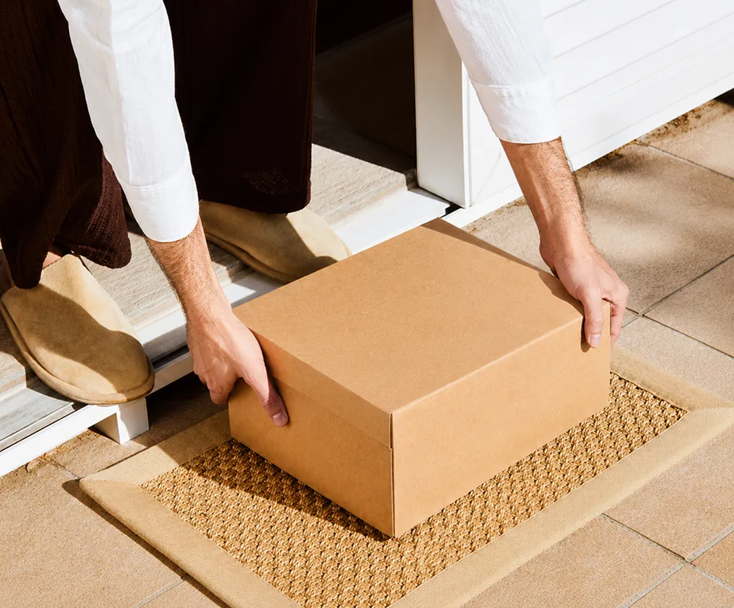 Person placing a cardboard box on a woven doormat outside a doorstep.