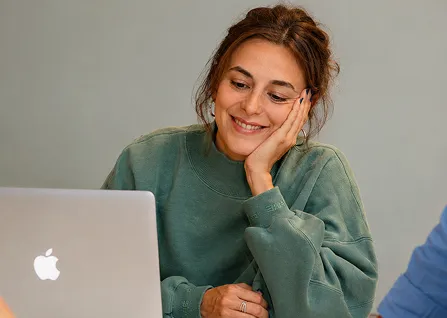Smiling woman in green sweatshirt looking at Apple laptop screen with hand on her cheek.