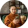 Person sitting indoors holding a tan ceramic mug and looking at it thoughtfully.