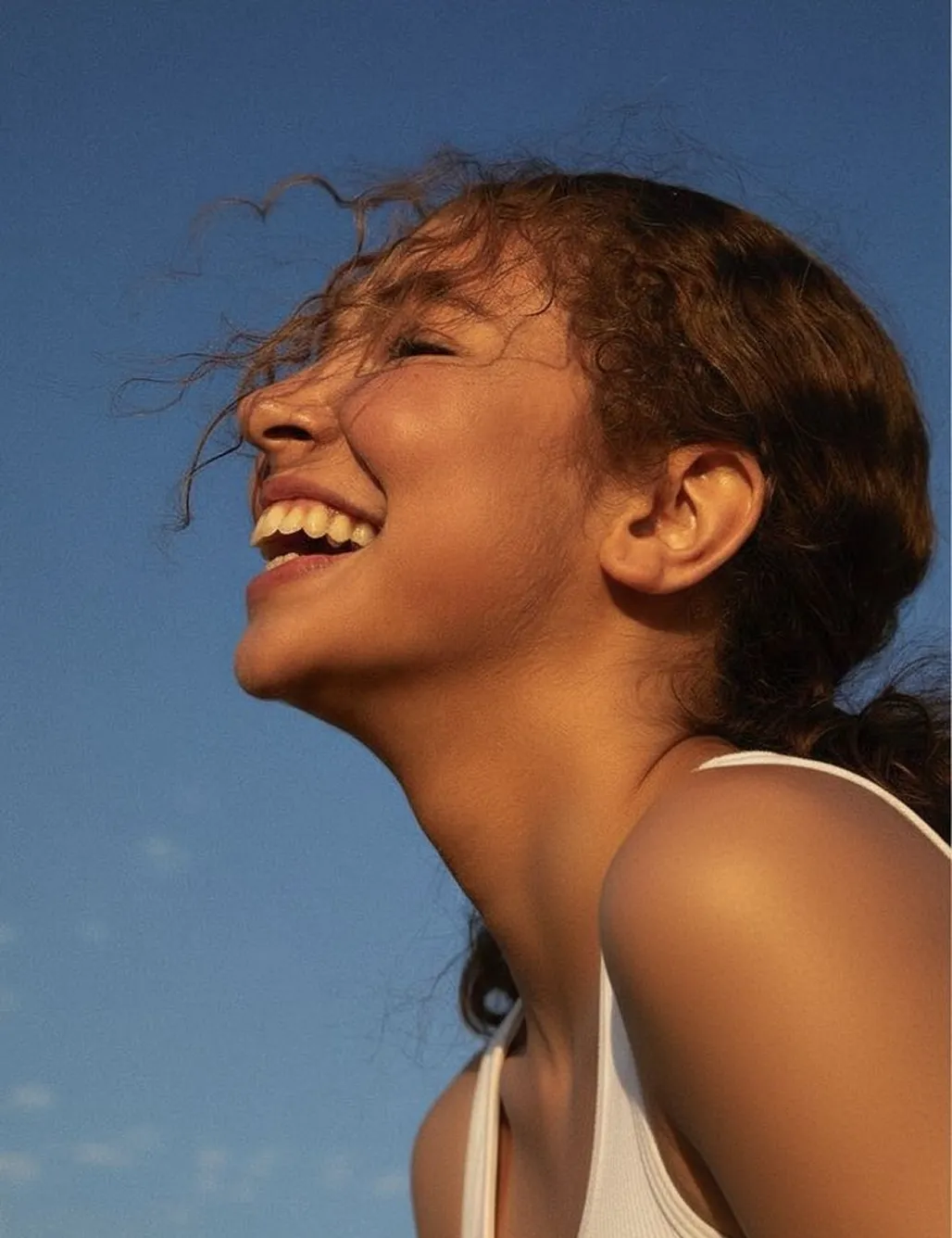 Side profile of a woman with curly hair laughing joyfully against a clear blue sky.