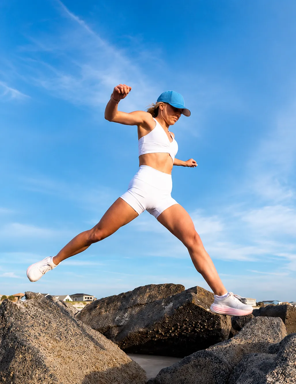 Athletic woman in white sportswear and blue cap jumping over large rocks under blue sky.