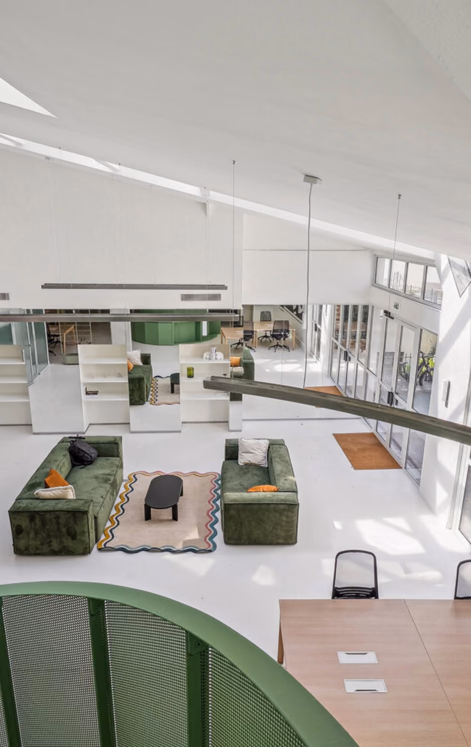 Vue en plongée d'un salon moderne avec deux canapés verts face à face, un petit tapis coloré central et une table basse noire, un grand espace lumineux avec de grandes fenêtres et un mur en verre, des luminaires suspendus, et une rambarde verte en métal au premier plan.