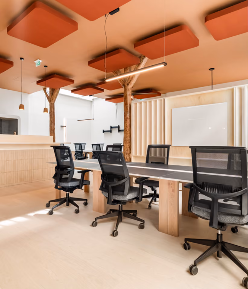 Espace de bureau moderne avec une grande table rectangulaire en bois clair entourée de chaises de bureau noires à roulettes, plafond avec panneaux acoustiques orange suspendus et piliers en bois naturel apparents.