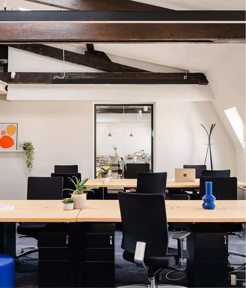 Bureau moderne avec plusieurs chaises de bureau noires autour de tables en bois clair, des pots de plantes vertes décorent les tables, poutres en bois sombres visibles au plafond, et une grande fenêtre intérieure reflète une autre salle.
