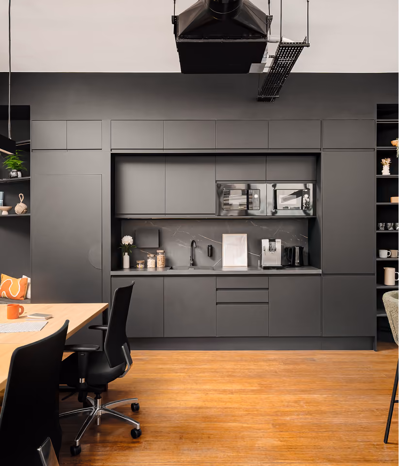 Cuisine moderne intégrée dans un bureau avec des armoires noires, un évier, un micro-ondes, une machine à café, des étagères ouvertes avec décorations, une table en bois clair avec chaises de bureau noires sur un sol en bois clair.