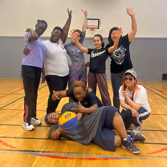 Groupe de huit personnes souriantes dans un gymnase sur un court de basket, certaines debout avec les bras levés, d'autres assises ou allongées tenant un ballon de basket jaune et bleu.