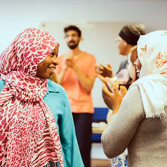Deux femmes portant des hijabs discutent joyeusement en premier plan, tandis que trois personnes floues échangent en arrière-plan dans un environnement intérieur lumineux.