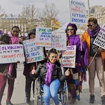 Un groupe de femmes portant des vêtements colorés et des écharpes violettes posent ensemble lors d'une manifestation, tenant des pancartes avec des slogans sur la lutte contre la violence et pour les droits des femmes.
