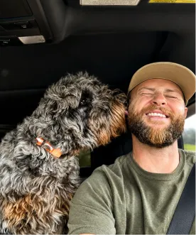 A man wearing a cap smiles as a large, curly-haired mountain doodle dog licks his ear inside a car.