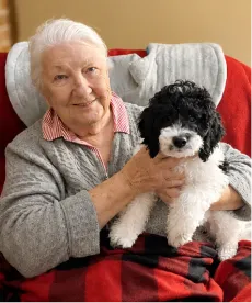 Older lady sitting in a chair, smiling and holding a small black and white dog on her lap.