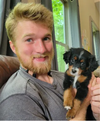 A man with a beard sits indoors and smiles holding a small black and brown puppy.
