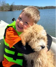 A young boy in a life jacket hugs a fluffy mountain doodle dog while sitting in a boat on a lake.