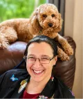 A happy owner with glasses smiles while sitting on a brown chair. A curly-haired dog lies on top of the chair behind them, looking at the camera.